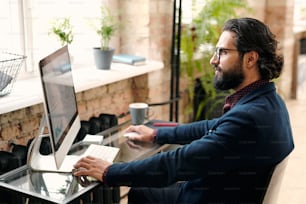 Serious young bearded broker or economist in formalwear sitting by table in front of computer monitor and studying financial data on screen