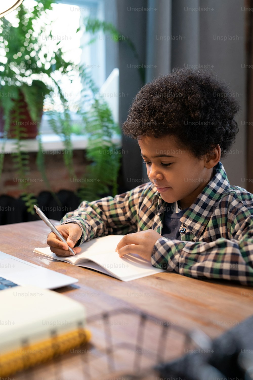 Süßer kleiner afrikanischer Schuljunge mit Stift, der Notizen auf einer Seite des Heftes macht, während er am Tisch vor dem Laptop sitzt und Hausaufgaben ausführt