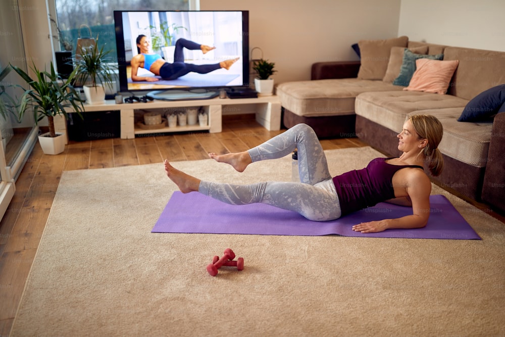 Atleta femenina practicando sus abdominales inferiores mientras sigue la clase de ejercicios en un televisor en la sala de estar.