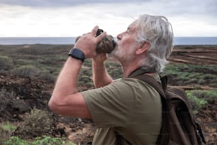 Side view of senior man with white beard enjoying the hike in arid landscape, standing and drinking at water bottle. Horizon over sea water