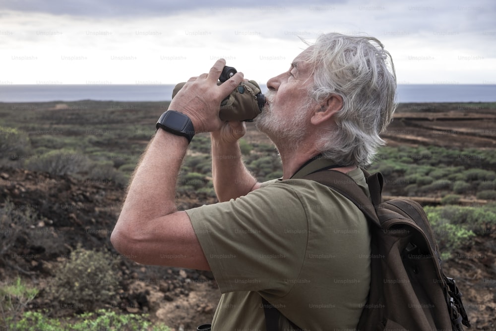 Side view of senior man with white beard enjoying the hike in arid landscape, standing and drinking at water bottle. Horizon over sea water