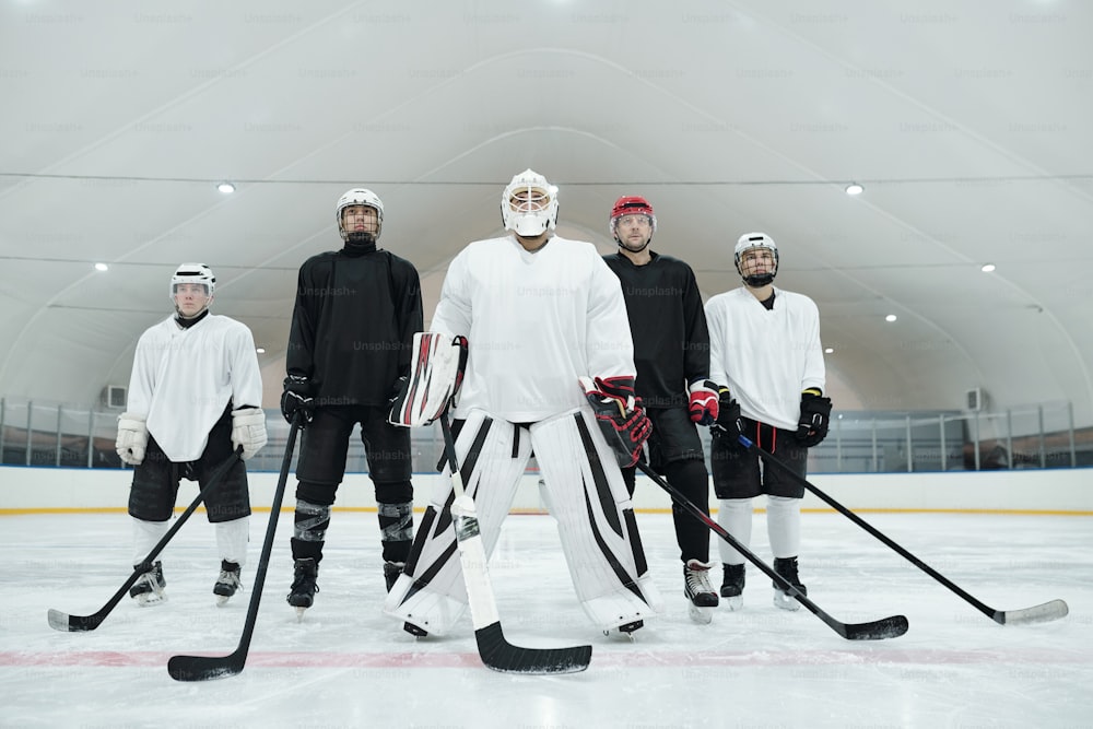 Vários jogadores de hóquei e seu treinador em uniforme esportivo, luvas, patins e capacetes em pé na pista de gelo e segurando bastões na frente de si mesmos