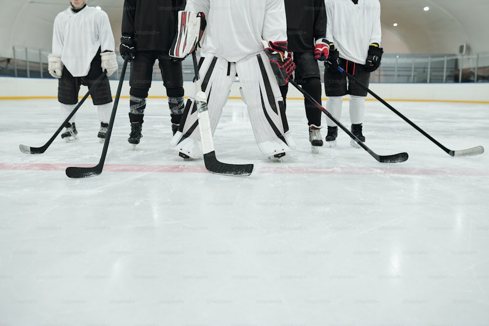 Unterer Teil mehrerer Hockeyspieler und ihres Trainers in Sportuniform, Handschuhen und Schlittschuhen, die auf der Eisbahn des Stadions stehen und Stöcke halten