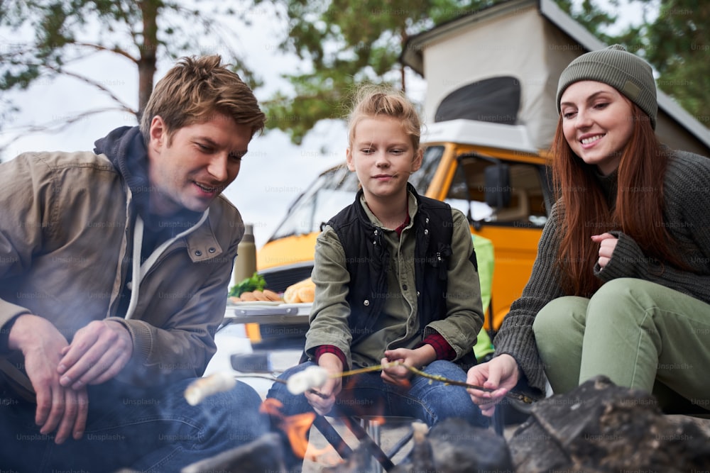 Campfire outdoor fun. Happy young family at camp or picnic, enjoy roasting marshmallow at fire, sitting near bonfires in a cozy wood, warming and talking