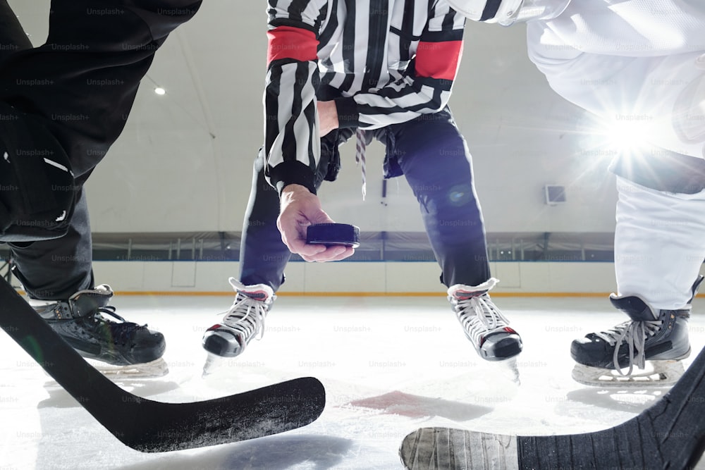 Árbitro de hockey con disco parado en la pista de hielo entre dos jugadores con palos esperando el momento para ser el primero en dispararlo durante el entrenamiento