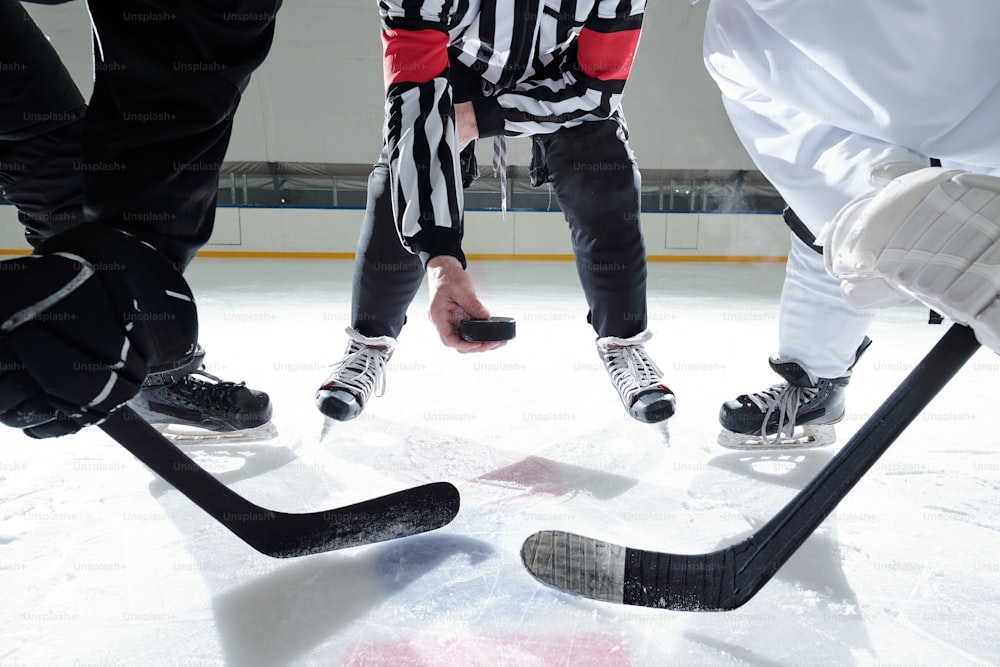 Árbitro de hockey con disco parado en la pista de hielo con dos rivales con palos a su derecha e izquierda esperando el momento para dispararlo