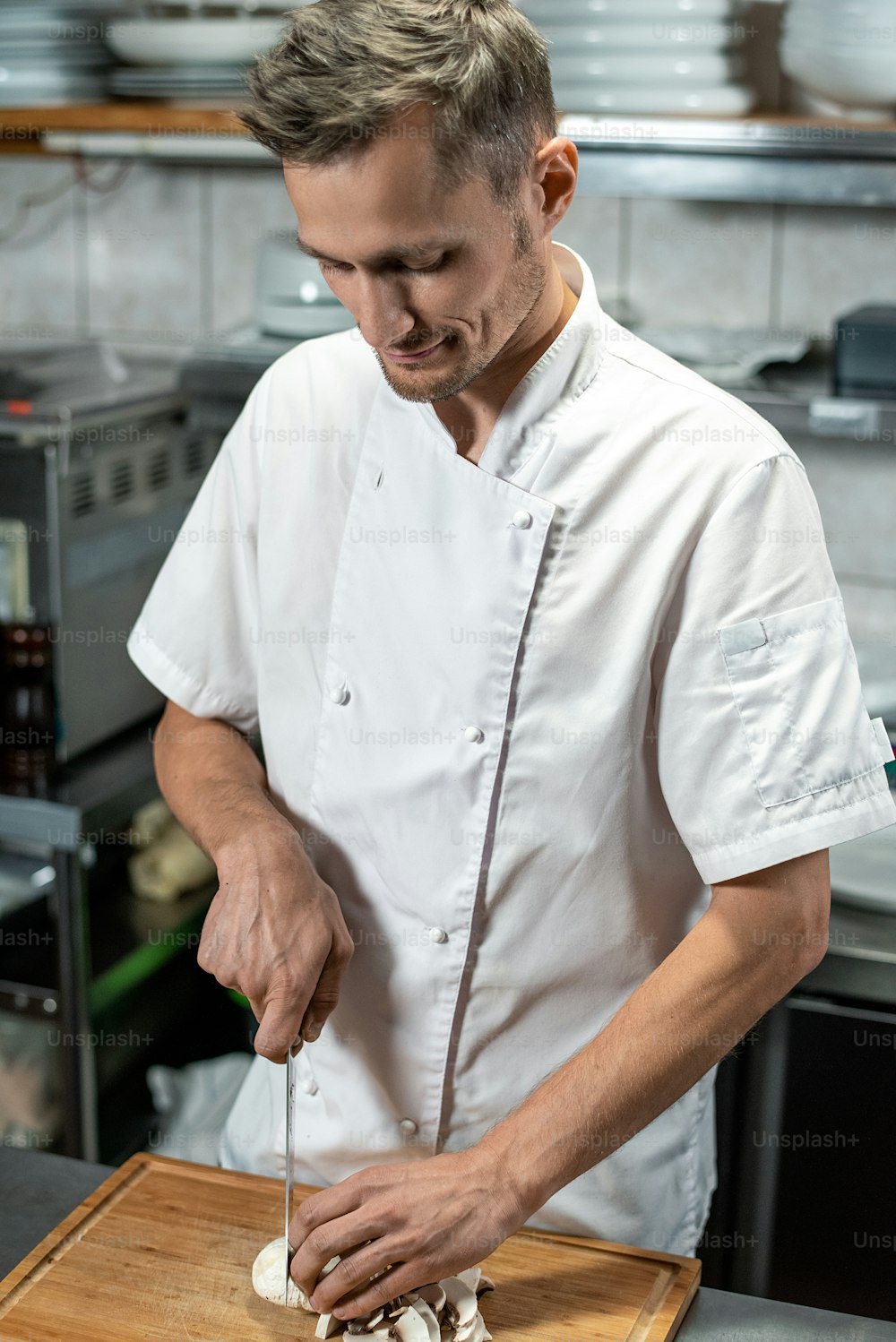 Young male chef of modern restaurant in white uniform chopping mushrooms for vegetable garnish while standing by table in the kitchen