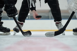 Hand of referee holding puck over ice rink with two players with sticks standing around and waiting for moment to shoot it