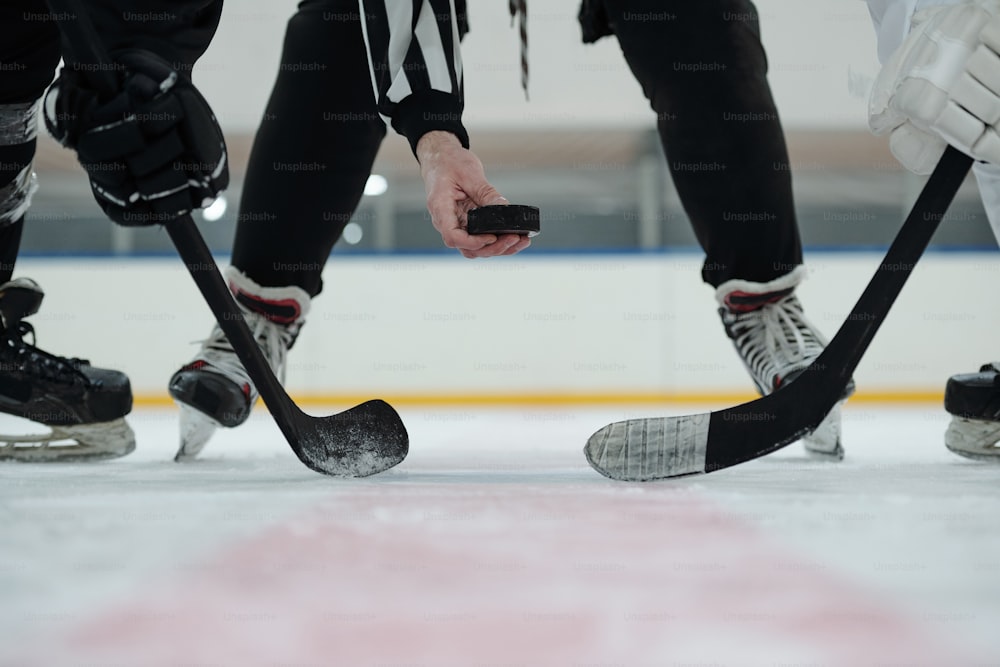 Mano del árbitro sosteniendo el disco sobre la pista de hielo con dos jugadores con palos parados alrededor y esperando el momento para dispararlo