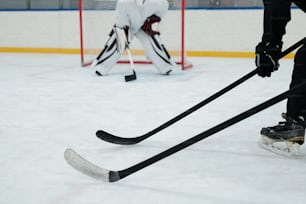 Bastoni da hockey tenuti da due giocatori in pattini, guanti e uniforme sportiva sullo sfondo del portiere che si prepara a prendere il disco
