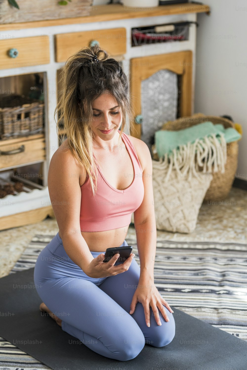 Aufnahme einer Fitnessfrau, die auf einer Yogamatte sitzt und ein Mobiltelefon benutzt. Fitte junge Frau, die Handy benutzt, während sie zu Hause Sport treibt. Online-Unterricht Sportliche Menschen mit gesundem Lebensstil
