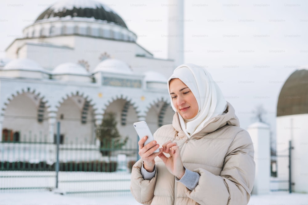 Bella giovane donna musulmana sorridente in foulard in abiti leggeri usando il cellulare sullo sfondo della moschea nella stagione invernale
