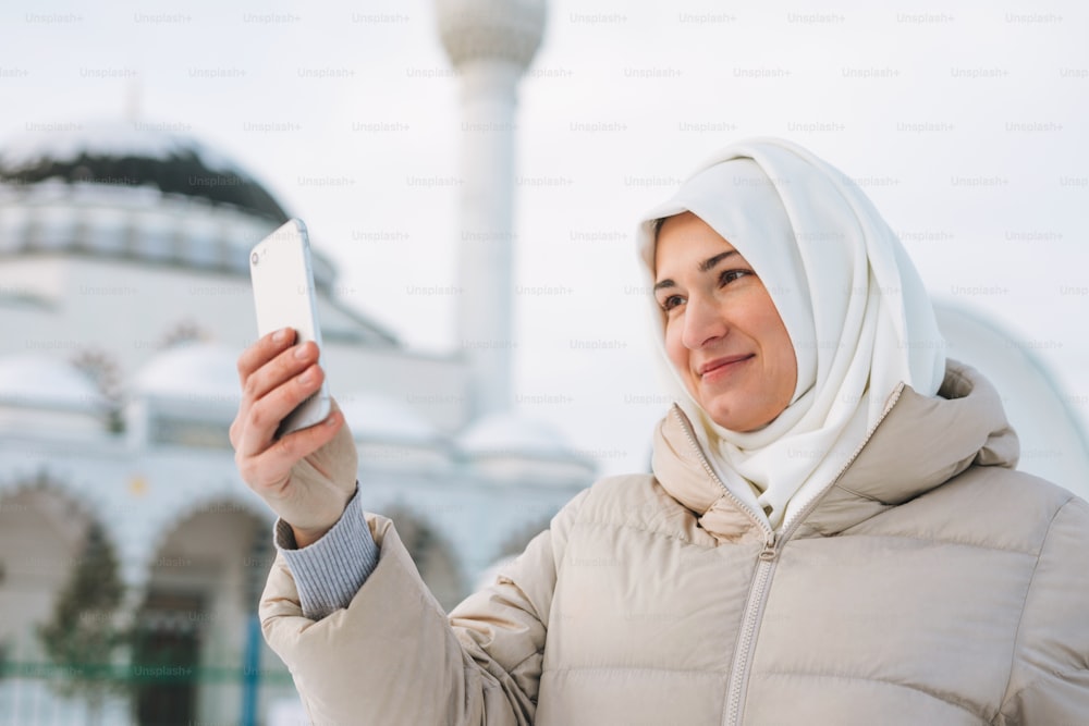 Beautiful smiling young Muslim woman in headscarf in light clothing takes selfie against the background of mosque in winter season