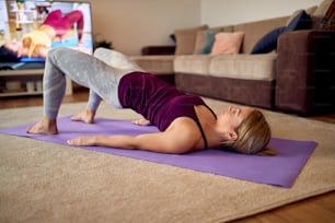 Dedicated sportswoman lifting her hips and doing glute bridge exercise while following instructions of her fitness instructor on a TV at home.