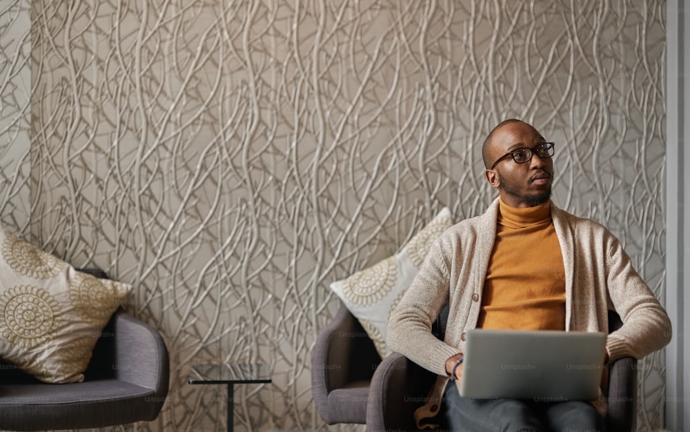 젊은 동기 부여 흑인 아프리카 사업가가 사무실 로비에 앉아 노트북으로 작업 중이며 복사 공간이 있는 거리를 바라보는 와이드 샷