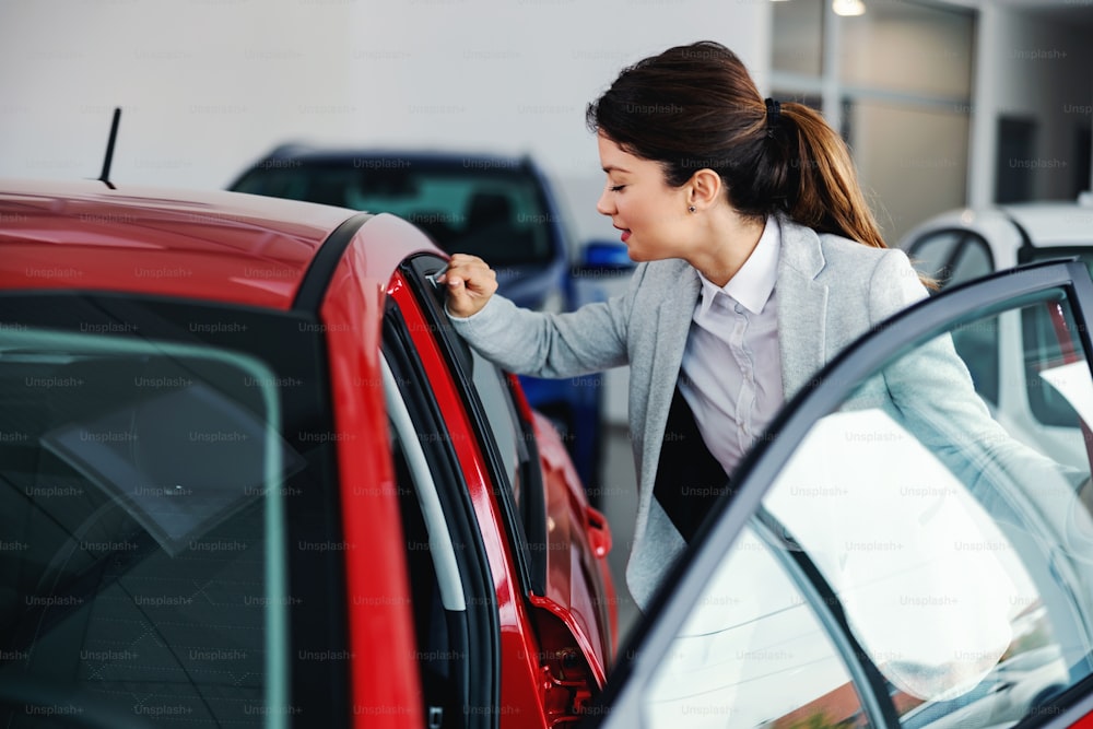Young tidy female car seller standing in car salon and rubbing car window with her sleeve.