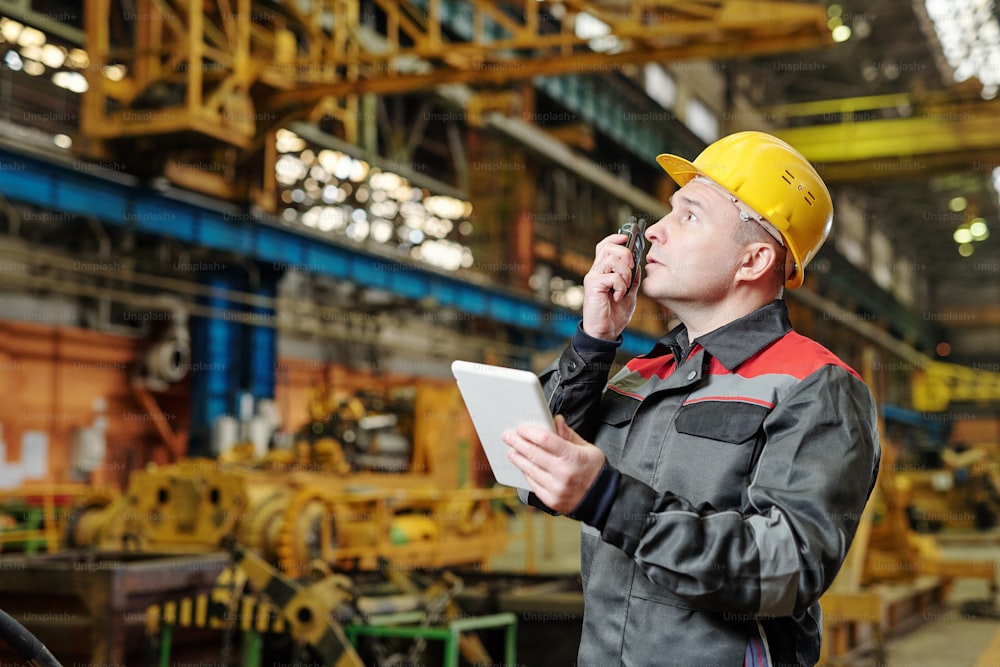 Mature foreman in helmet communicating on a wireless radio with his colleagues and using tablet pc in the plant