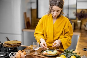 Young woman dressed in yellow bathrobe preparing eggs for breakfast, putting greens on a plate on the kitchen at home