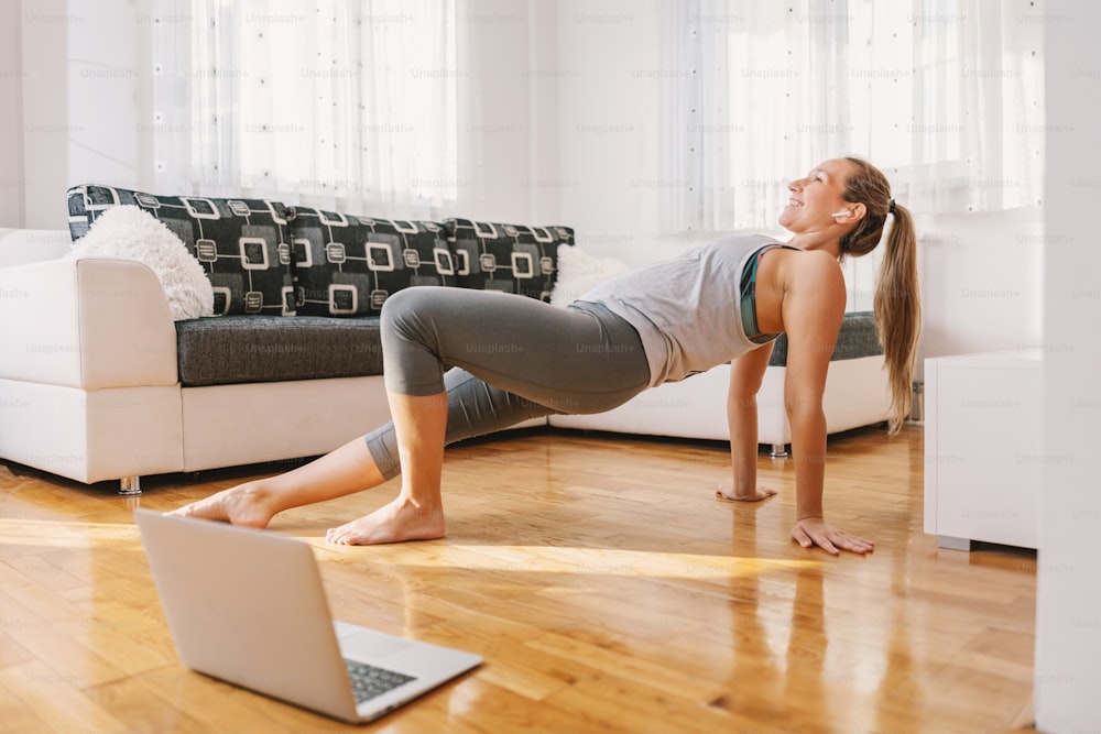Smiling muscular fitness instructor sitting at home on the floor and showing stretching exercises. She is having online class over laptop.