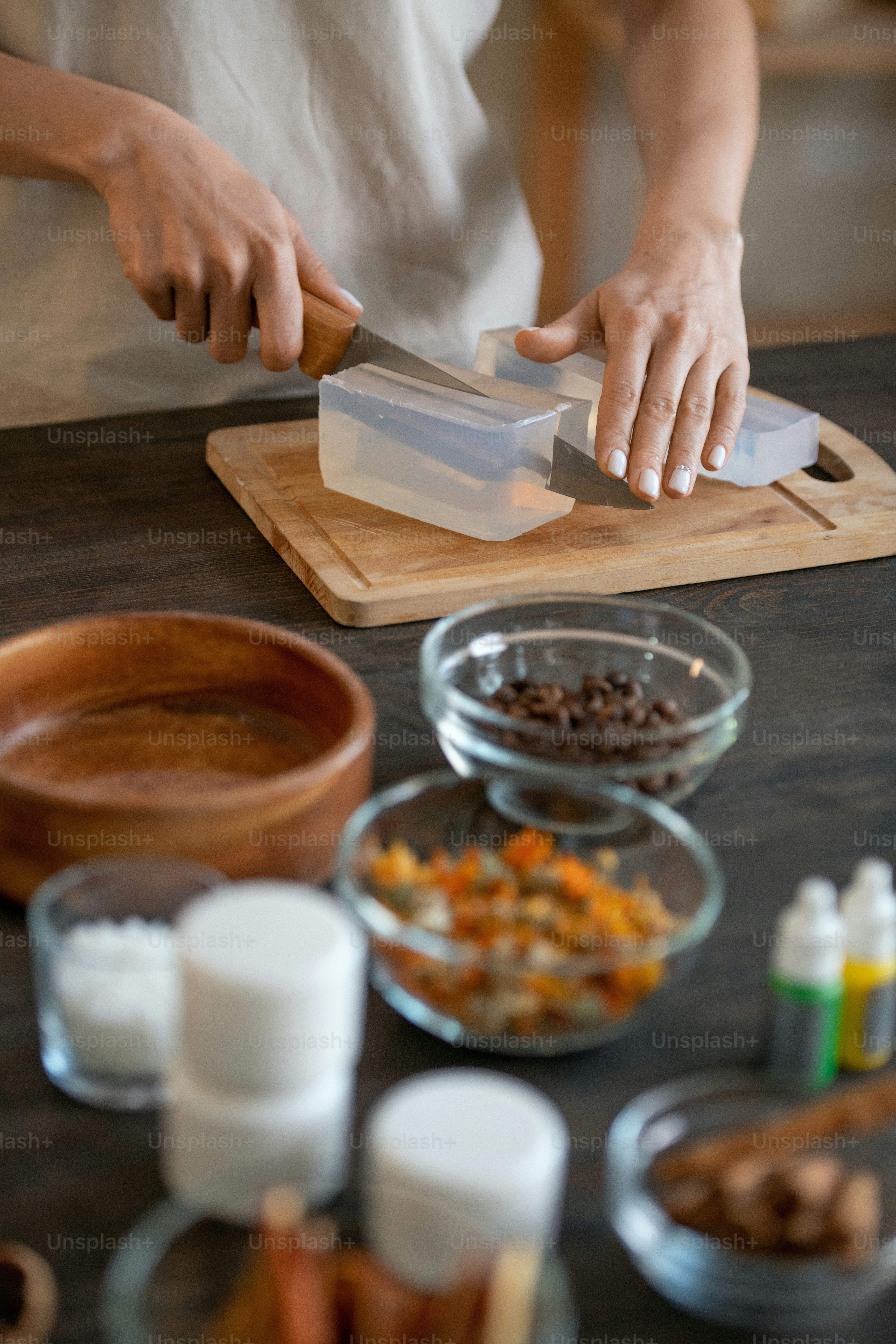 Manos de una mujer joven de pie junto a la mesa y cortando masa de jabón duro en un tablero de madera mientras hace productos cosméticos naturales en casa