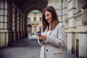 Smiling young brunette standing on the street and using phone for texting or hanging on social media.