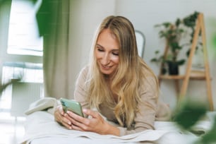 Beautiful blonde smiling young woman using mobile on bed in bright room at home