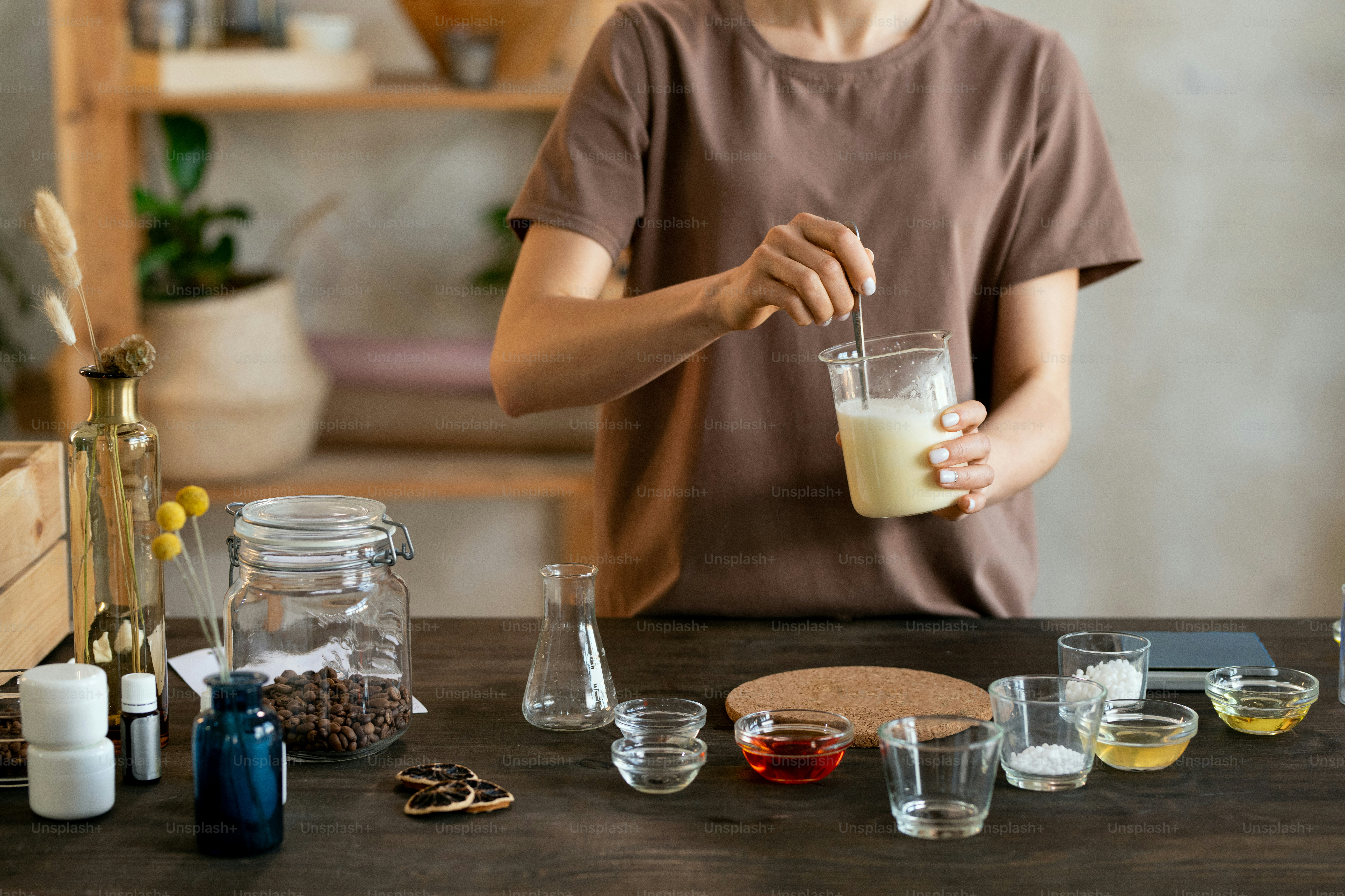 Wanita muda memegang barang pecah belah dengan massa sabun leleh di atas meja dengan barang pecah belah sambil membuat campuran untuk produk kosmetik buatan tangan alami