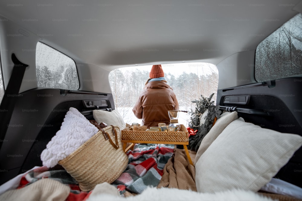 Donna seduta nel bagagliaio dell'auto vicino a una pineta, in viaggio in auto durante le vacanze invernali. Vista dal retro. Foto di alta qualità