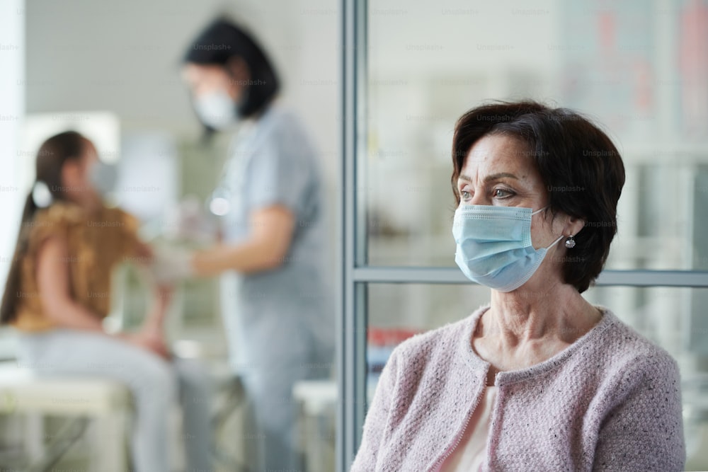 Mature woman in protective mask sitting in clinics and waiting for her turn to have vaccination against nurse making injection to little girl