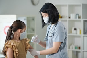 Young brunette nurse of Asian ethnicity in uniform and protective mask making injection to little girl sitting in medical office in clinics