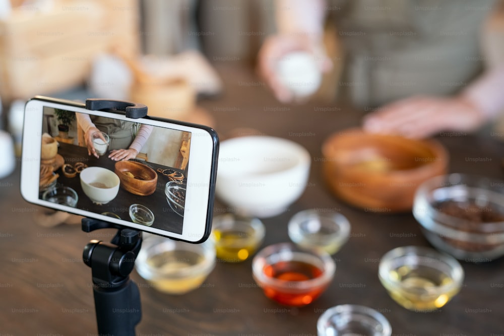 Teléfono inteligente en el soporte de la cámara de pie frente a la mesa donde una mujer joven prepara los ingredientes para el jabón natural hecho a mano