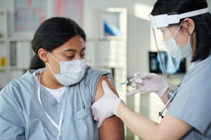 Médica contemporánea con uniforme y ropa de trabajo protectora inyectando a su colega mestiza durante la vacunación contra el covid