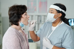 Mature woman with open mouth sitting in front of young nurse testing her for covid with oral swab in medical office of contemporary clinics
