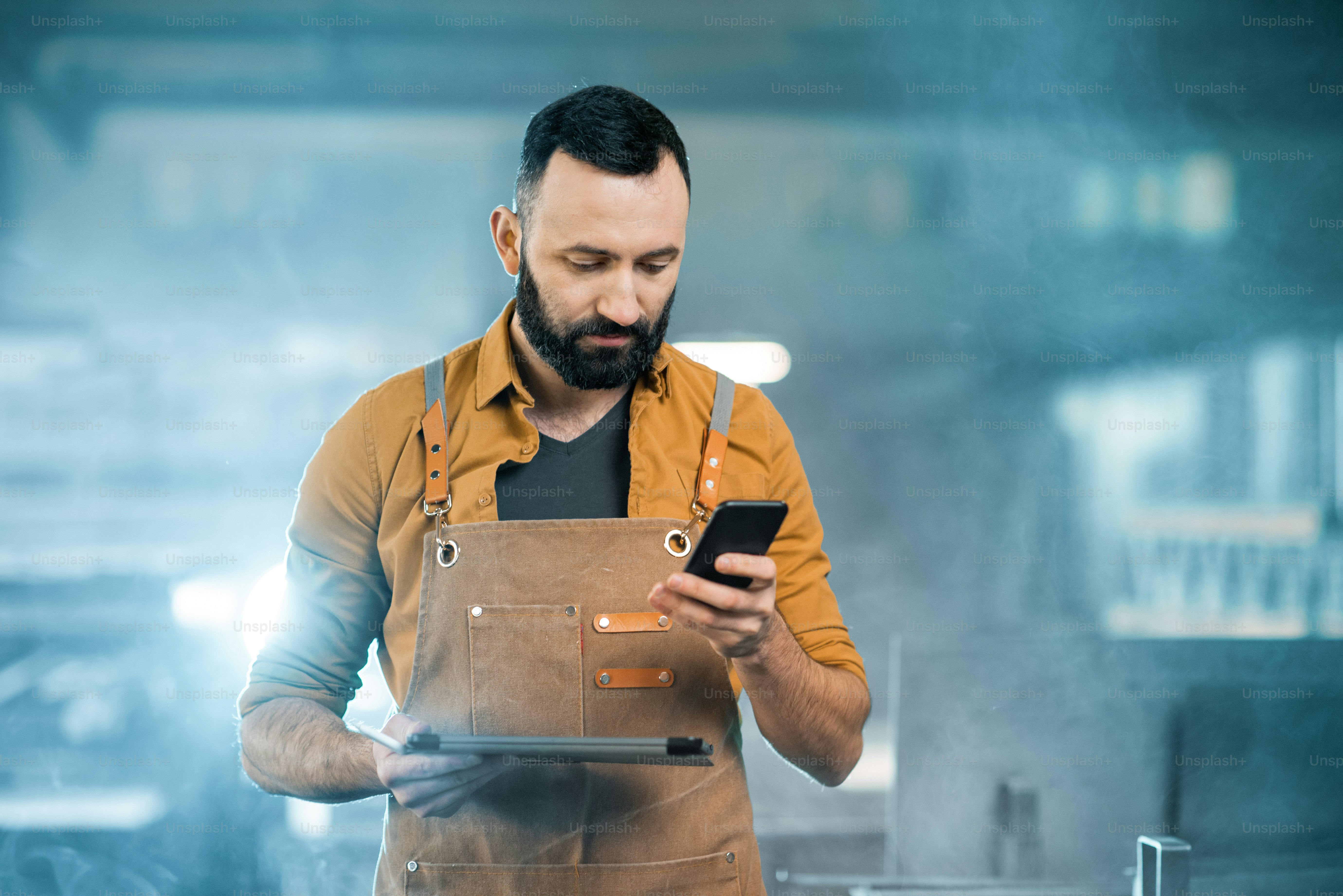A bearded man in a brown apron holding a smartphone and a tablet