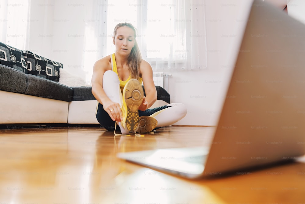 Fitness instructor sitting on te floor and tying shoelace. In front of her is laptop. She is preparing to have online class.