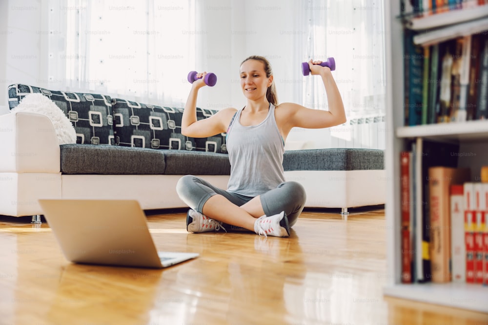 Strong sportswoman sitting on the floor at home and lifting dumbbells. She is following online tutorials.