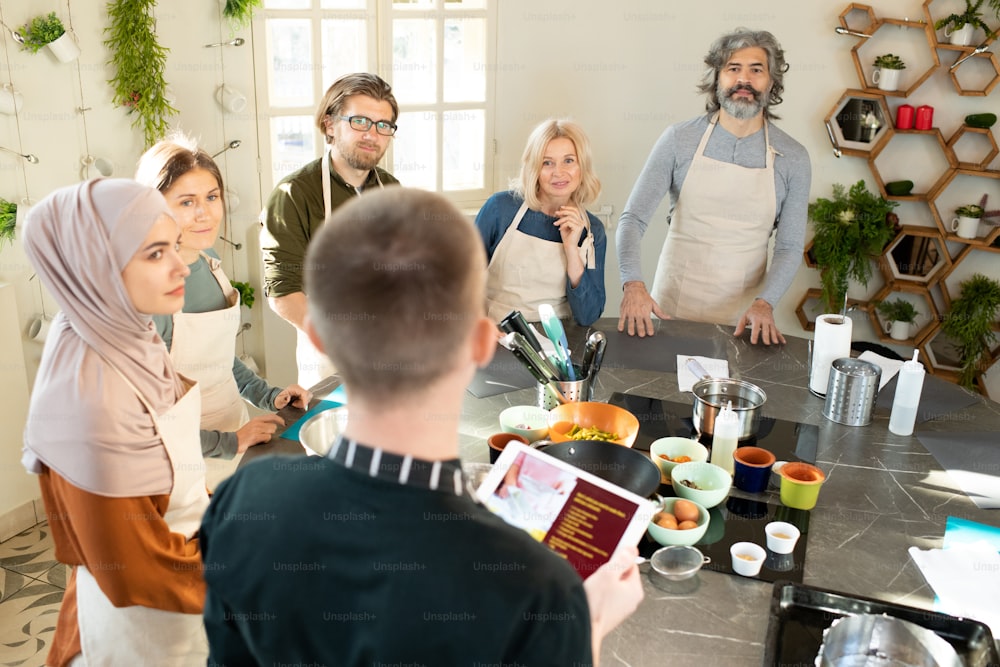 Group of serious people in aprons looking at male coach with digital tablet and listening to him during master class while standing around table