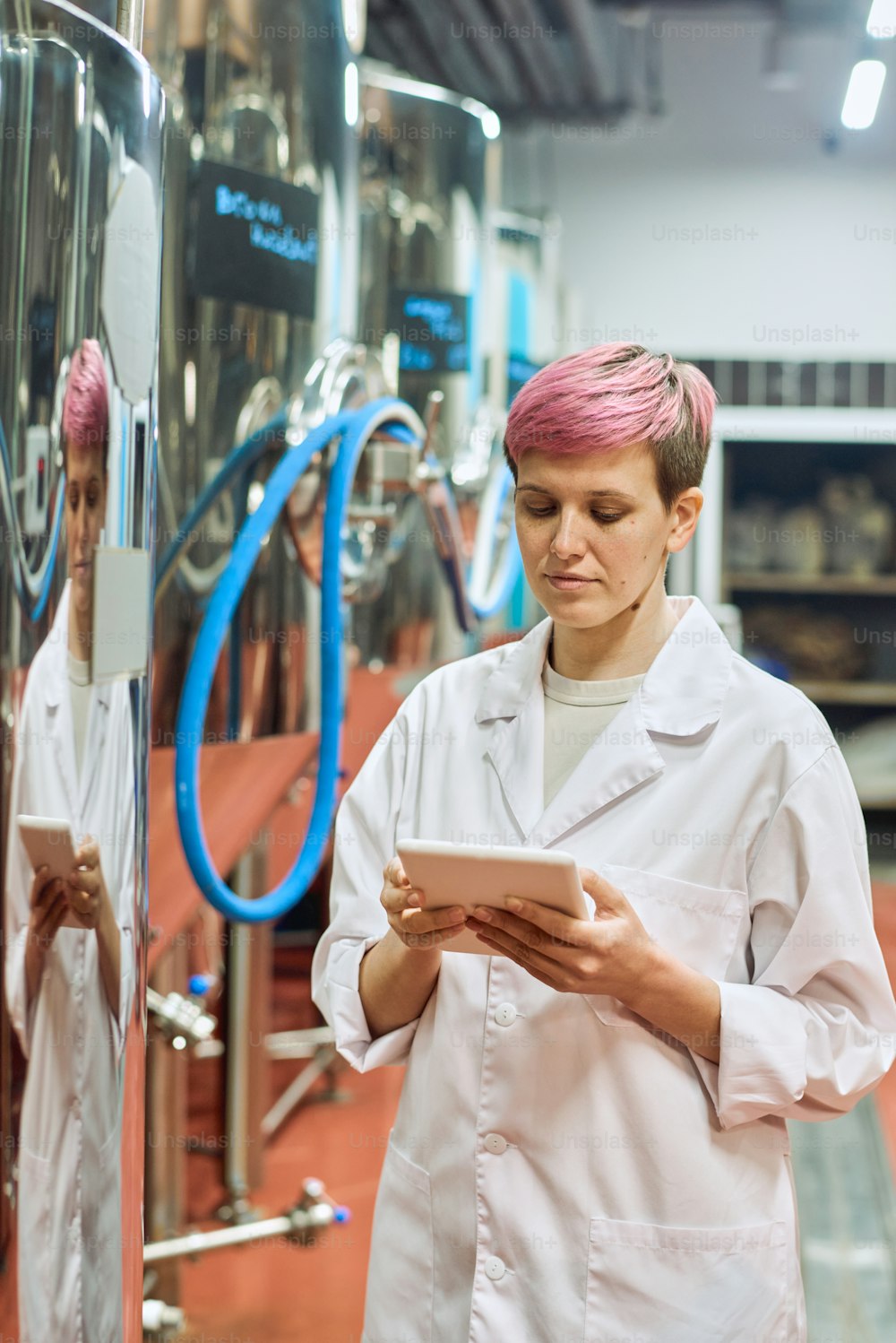 Giovane esperta di birrificio femminile in camice bianco che esamina i dati nella tavoletta digitale mentre si trova accanto a nuove attrezzature per la produzione di birra in pianta