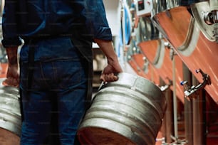 Vue arrière d’un ouvrier d’une usine de production de bière moderne en tenue de travail portant deux citernes tout en se déplaçant le long d’une rangée d’énormes réservoirs en acier