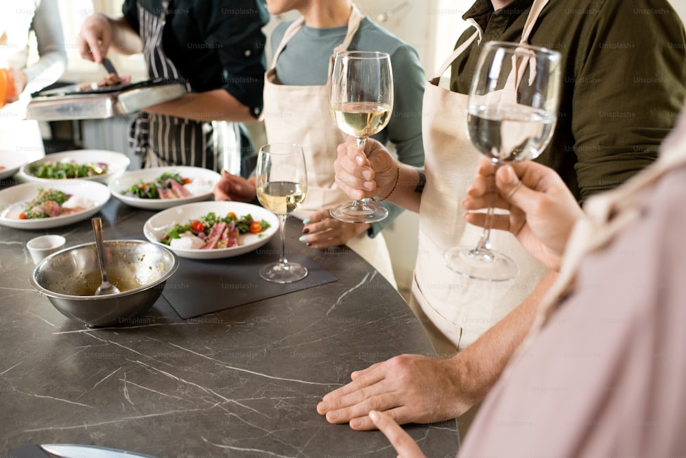 Gruppo di persone in grembiuli che brindano con flûte di champagne o vino bianco sopra il grande tavolo della cucina con il cibo fatto in casa appetitoso servito