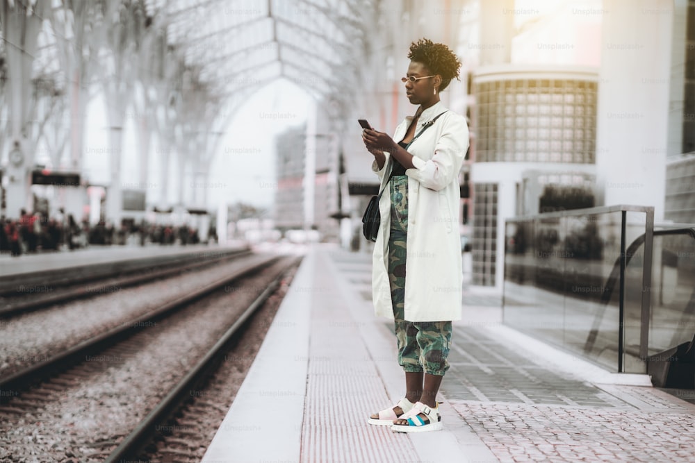 Une jeune femme africaine aux cheveux bouclés, portant des lunettes de soleil, un trench blanc et un pantalon de camouflage, téléphone alors qu’elle se tient sur le quai d’un dépôt de gare et attend que le train circule