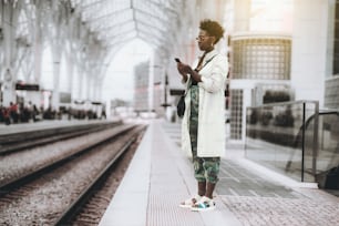 サングラスをかけ、白いトレンチ、迷彩柄のズボンを履いた若い派手な巻き毛のアフリカ人女性が、駅のプラットホームに立って列車を待っている