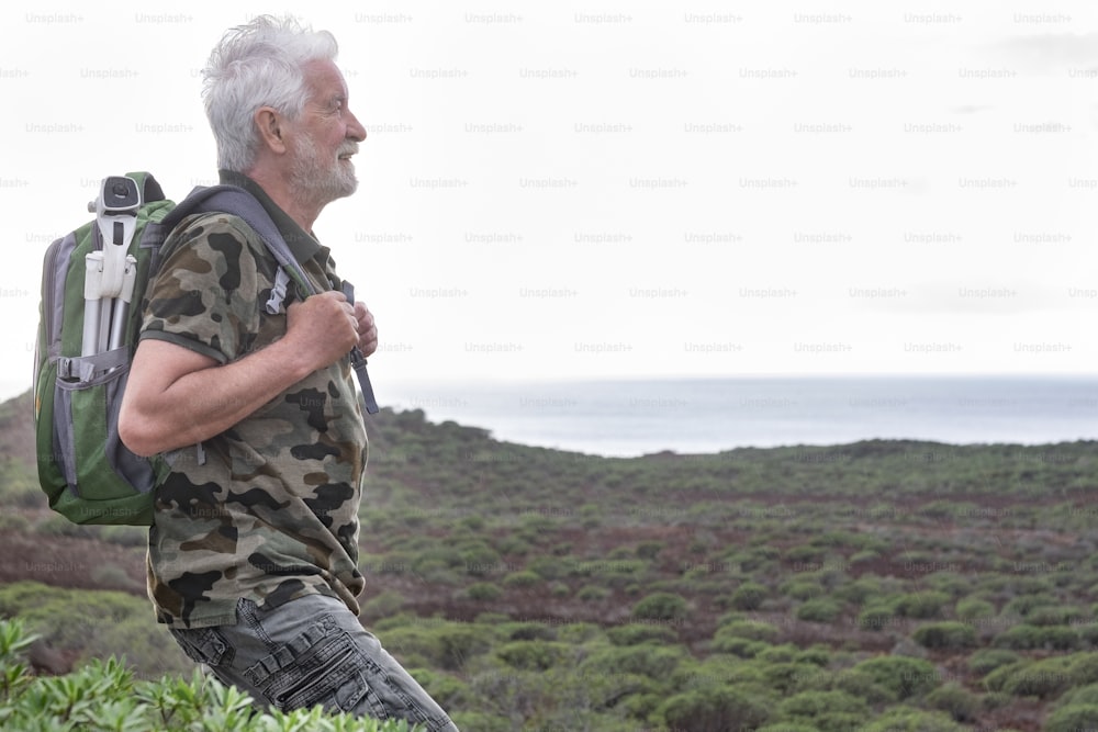 A smiling senior adult man with backpack on shoulders enjoying outdoors excursion between green bushes and sea. A white-haired elderly people in healthy activity