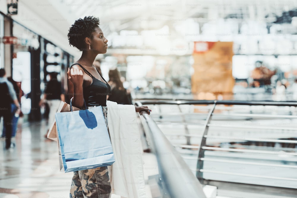 A beautiful young elegant black woman is standing next to the railing of a modern shopping mall with disposable paper bags with purchases in her hand after shopping; a copy space place on the right