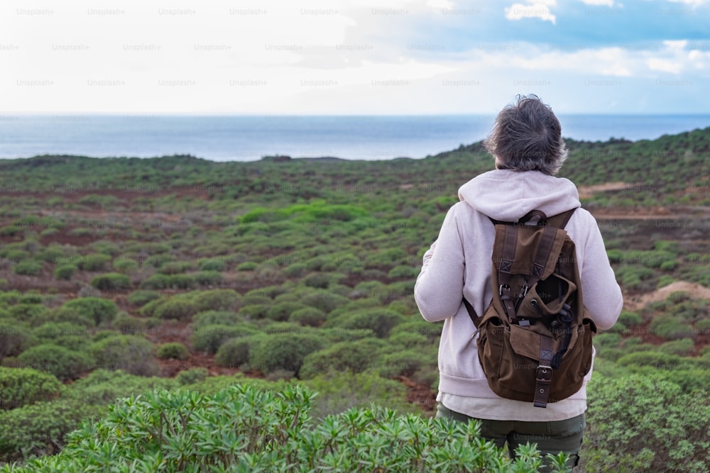 Vista trasera de una mujer mayor disfrutando de una excursión al aire libre entre arbustos verdes y el mar. Ancianos de pelo gris con mochila mirando al horizonte