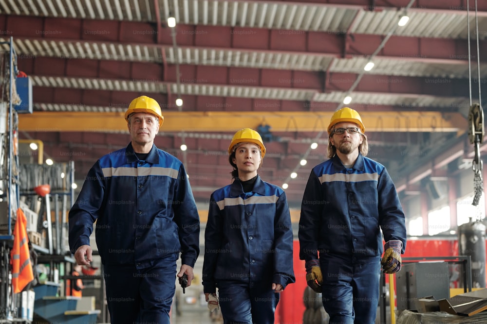 Gruppo di operai o ingegneri maschi e femmine di successo della fabbrica contemporanea che si muove lungo l'officina con l'attrezzatura industriale