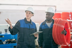 Young contemporary female worker of modern plant using tablet and showing her African male colleague new industrial machine