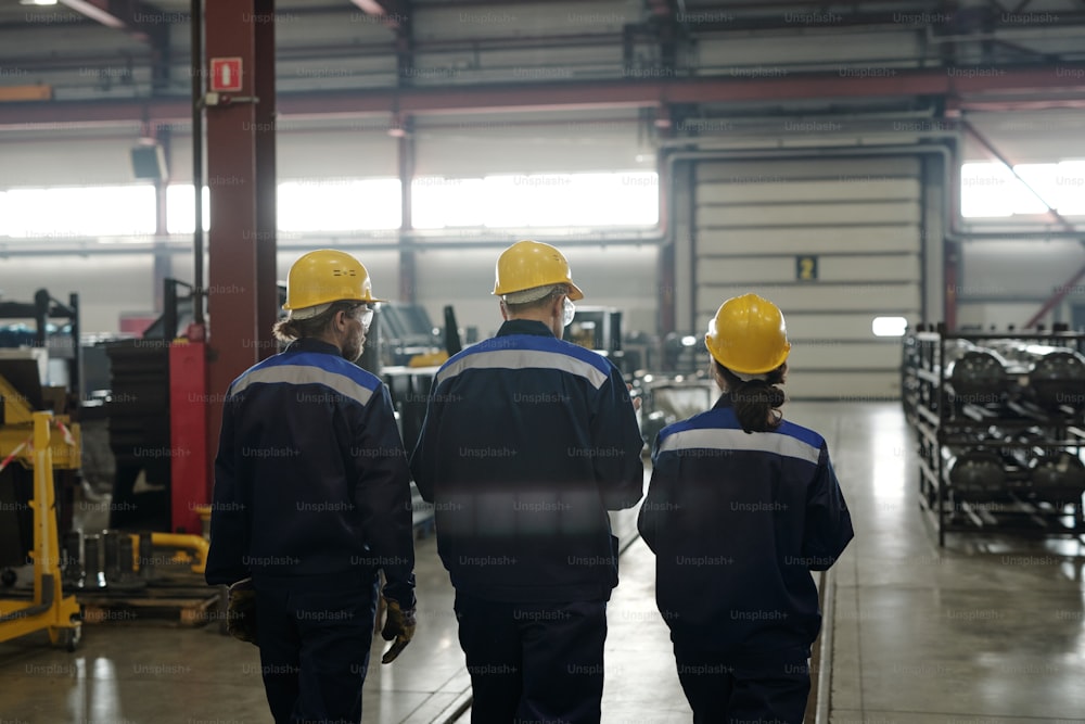 Vista posteriore del gruppo di operai o ingegneri di grande fabbrica contemporanea che si muove lungo l'officina con l'attrezzatura industriale e parla