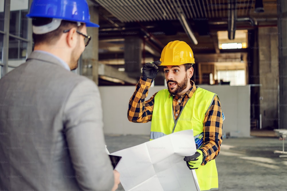 Construction worker holding blueprints and talking to his supervisor while standing at construction site.