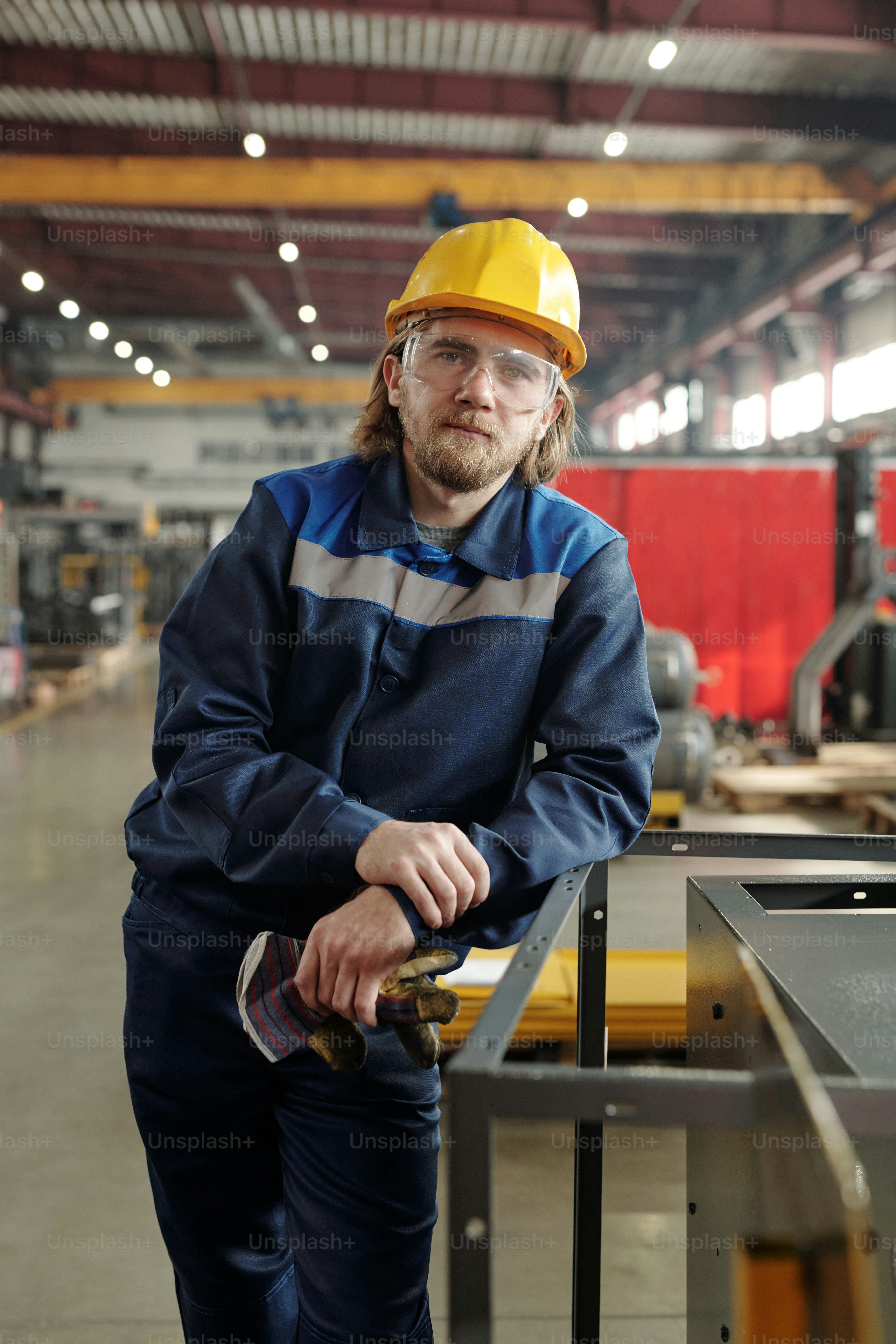 Junger bärtiger männlicher Arbeiter eines großen Betriebs oder einer großen Fabrik oder Vorarbeiter in Arbeitskleidung und Schutzhelm, der sich an Industrieanlagen lehnt